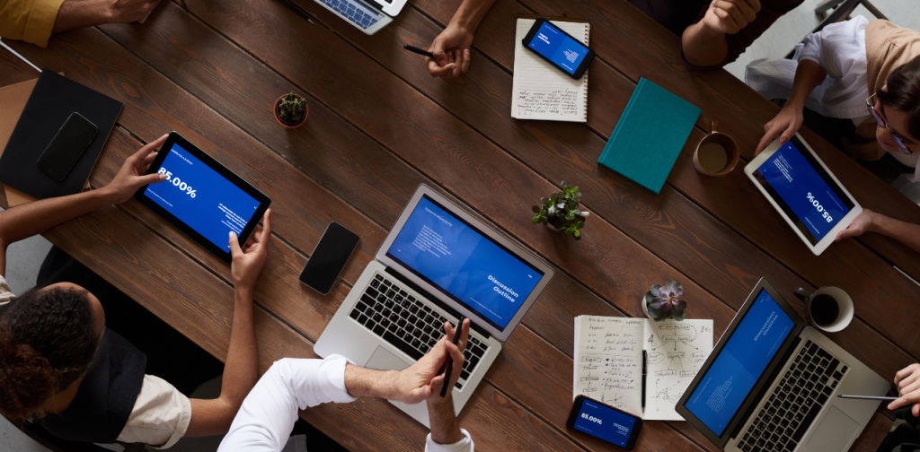 mehrere Personen sitzen rund um einen Tisch, vor sich Tablets und Laptops